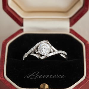 May include: A silver engagement ring with a large, round, clear gemstone as the centerpiece. The band has a unique, swirling design with smaller accent stones. The ring is in a red and black jewellery box, with the word "Lumea" printed on the box.