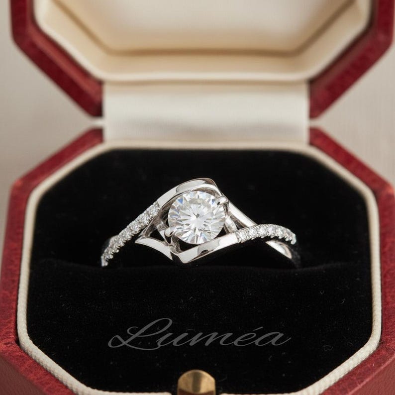 May include: A silver engagement ring with a large, round, clear gemstone as the centerpiece. The band has a unique, swirling design with smaller accent stones. The ring is in a red and black jewellery box, with the word "Lumea" printed on the box.