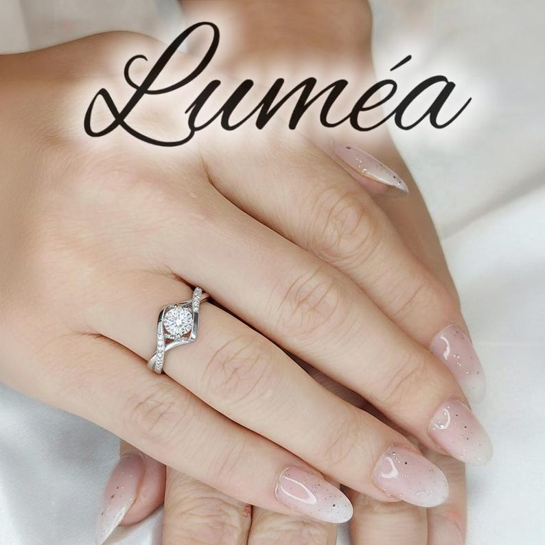 May include: A silver ring featuring a central clear gemstone and smaller surrounding stones. The ring is displayed on a hand with manicured nails. The word "Lumea" is written in cursive at the top of the image.