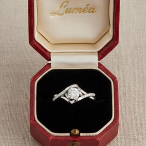 May include: A diamond engagement ring presented in a red octagonal box. The ring has a large, round, clear diamond set in a swirling, silver band. The box interior is black velvet, with the word "Lumea" in gold script on the lid.