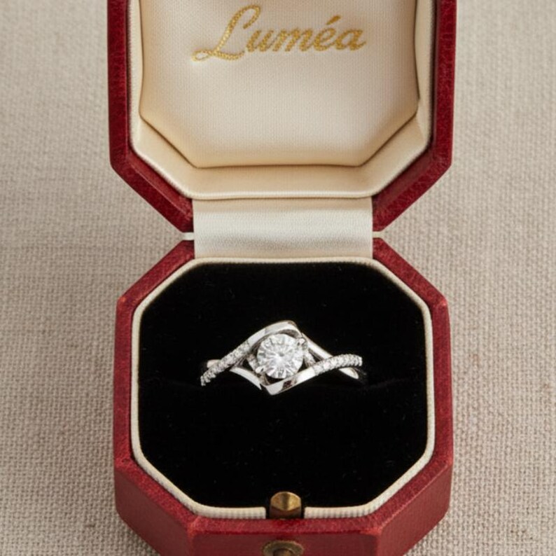 May include: A diamond engagement ring presented in a red octagonal box. The ring has a large, round, clear diamond set in a swirling, silver band. The box interior is black velvet, with the word "Lumea" in gold script on the lid.