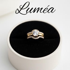May include: A gold engagement ring set with a large, round, clear gemstone and smaller accent stones. The set includes a curved band and a matching band, all displayed in a black ring box. The word "Lumea" is written above the rings.