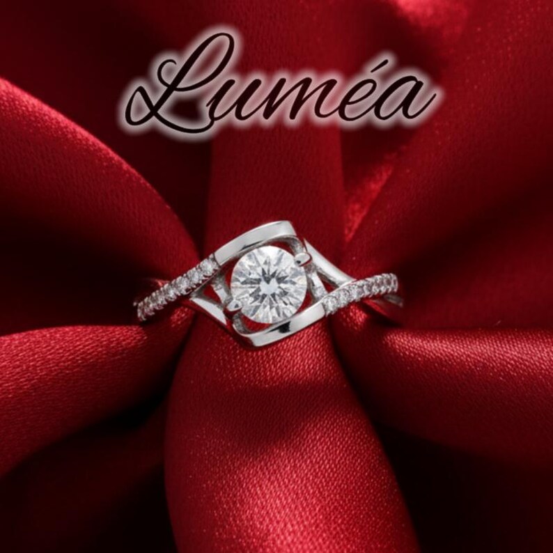 May include: A silver ring with a central, clear, round gemstone and smaller surrounding stones. The ring features a distinctive, flowing design, set against a red satin backdrop. The word "Lumea" is written in cursive at the top.
