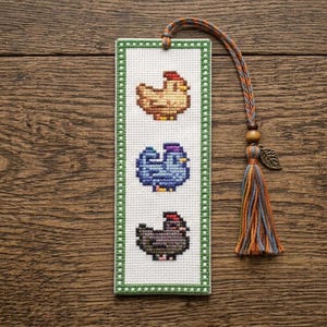 May include: A rectangular cross-stitch bookmark on white fabric with a green border, featuring three stacked chicken motifs in brown, blue, and dark gray. A braided cord with a wooden bead, a metal leaf charm, and a multi-colored tassel is attached.