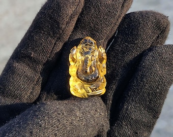 Puede incluir: Una figura tallada, translúcida, de color amarillo-naranja del dios hindú Ganesha. La escultura se sostiene en un guante oscuro, con detalles del dios con cabeza de elefante visibles. La figura es un colgante o una pequeña estatua.