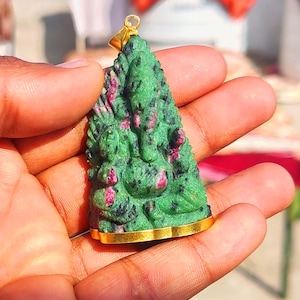 May include: A green and pink gemstone pendant in the shape of a deity, with a gold-colored bail and base. The pendant is held in a hand, showcasing the intricate details of the carved figure.