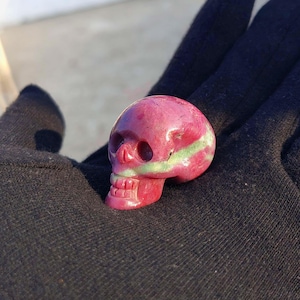 May include: A small, carved skull figurine in shades of pink and green. The skull has detailed features, including eye sockets, a nose cavity, and teeth. The figurine is held in a black gloved hand.