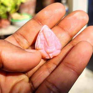 Puede incluir: Pequeña escultura de piedra rosa tallada de una deidad, sostenida en una mano. La escultura tiene detalles finos y una textura suave. El color es un rosa pastel suave.