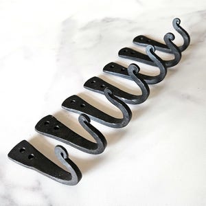 May include: A set of seven black metal hooks arranged diagonally across a white marble surface. Each hook has a flat, rectangular base with two holes and a curved, scrolled end. The hooks are made of a dark, textured metal.