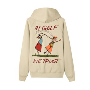 Könnte beinhalten: Beigefarbener Kapuzenpullover mit Kapuze und langen Ärmeln. Auf der Rückseite befindet sich eine Cartoon-Illustration von zwei Golfspielern mit dem Text "IN GOLF WE TRUST" in Rot und Schwarz. Das Design ist eine spielerische Interpretation von Golf.