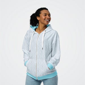 May include: A light grey zip-up hoodie with a delicate pattern. The hoodie has a light blue lining in the hood and cuffs, and white drawstrings. The model is wearing blue jeans.