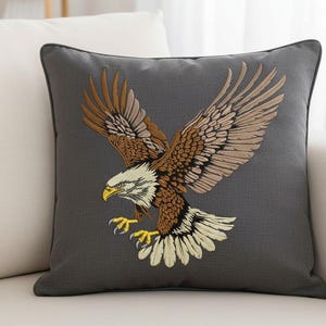 Puede incluir: Un cojín decorativo cuadrado gris con un águila bordada en vuelo. El águila tiene plumas marrones, beige y crema, una cabeza blanca y un pico y garras amarillas. El cojín tiene un borde gris oscuro.