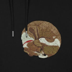 Puede incluir: Sudadera con capucha negra con un parche bordado de un zorro durmiendo. El zorro está representado en tonos marrones, blancos y rojos, con costuras detalladas. La sudadera tiene un cordón y ojales metálicos.