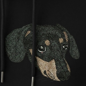 Puede incluir: Sudadera con capucha negra con un diseño de perro salchicha bordado. La cabeza del perro está detallada con hilo negro, marrón y beige, mostrando sus ojos y hocico. La sudadera tiene cordones.
