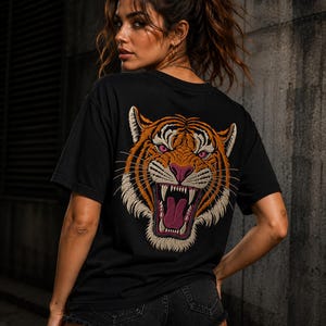 Puede incluir: Una camiseta negra de manga corta con un gráfico grande y detallado en la espalda de una cabeza de tigre rugiendo. El tigre está representado en tonos naranja, blanco y negro con ojos rosas, nariz rosa y la boca abierta mostrando dientes afilados.