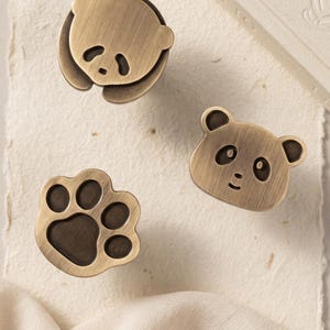 Peut inclure: Trois boutons de meuble de couleur bronze avec des motifs de panda et de patte. L'un est un panda couché, un autre un visage de panda et le troisième une empreinte de patte. Les boutons sont en métal et ont un aspect vintage.