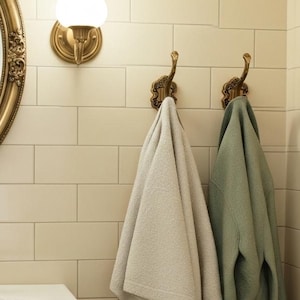 Peut inclure: Intérieur de salle de bain avec un miroir ovale doré, un lavabo blanc avec des robinets dorés et deux crochets décoratifs tenant un peignoir gris clair et un peignoir vert sauge. Murs carrelés crème.