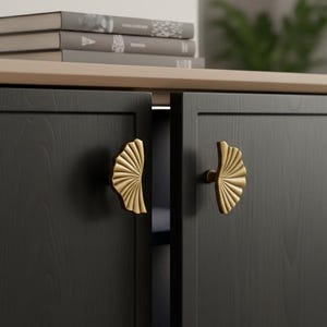 Peut inclure: Un meuble noir avec des poignées dorées en forme d'éventail. Le meuble a un dessus en bois clair et est placé sur un fond neutre. Des objets décoratifs sont visibles sur le dessus du meuble.