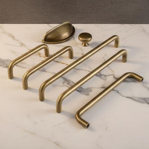 Può includere: Una collezione di ferramenta per mobili in ottone antico, tra cui diverse maniglie a barra, una maniglia a tazza e un pomello rotondo. L'hardware è disposto su una superficie di marmo bianco.