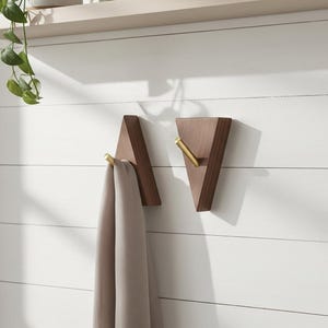 May include: Two wooden wall hooks with brass-colored hooks. The hooks are triangular in shape and mounted on a white, horizontal-slatted wall. One hook holds a beige scarf. A shelf with a plant is visible in the background.