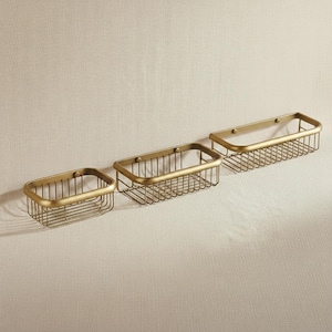 May include: Three rectangular, gold-coloured wire baskets of varying sizes are displayed against a neutral background. Each basket has a rounded top frame and is designed for wall mounting. The baskets are likely for bathroom or kitchen storage.