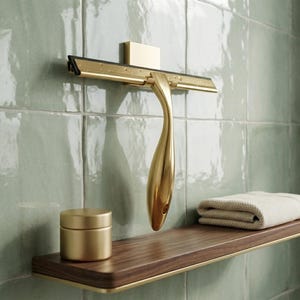 Könnte beinhalten: Ein goldfarbener Duschabzieher mit gebogenem Griff ist an einer grün gefliesten Wand montiert. Ein Holzregal mit goldenen Zierleisten hält ein kleines goldenes Glas und ein gefaltetes beiges Handtuch. Der Abzieher ist für die Reinigung von Duschtüren konzipiert.