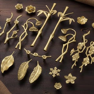 Op de afbeelding: Een assortiment gouden decoratieve hardware, waaronder ladegrepen, knoppen en decoratieve accenten. De ontwerpen bevatten bloemen-, blad- en bamboemotieven. De hardware is gerangschikt op een donker houten oppervlak.