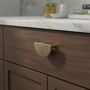 Peut inclure: Une poignée de meuble dorée, semi-circulaire, avec une surface texturée, est fixée à un tiroir de meuble brun foncé. La poignée est montée horizontalement et présente un design moderne. Le meuble fait partie d'un ensemble de cuisine ou de salle de bain.