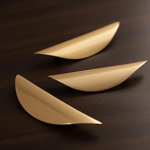 Peut inclure: Trois poignées de meubles dorées et incurvées disposées sur une surface marron foncé. Les poignées ont une finition lisse et mate et une forme de feuille, avec une extrémité pointue et une base plus large et arrondie.
