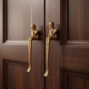 Può includere: Due maniglie dorate per armadi, ciascuna a forma di figura seduta con gambe lunghe e una mano che sostiene la testa. Le maniglie sono montate su un'anta di armadio in legno marrone scuro, creando un elemento di design unico e artistico.