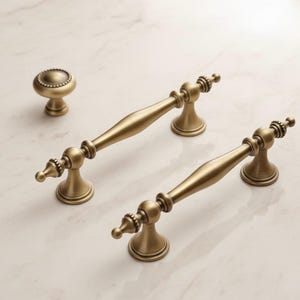 May include: Antique brass cabinet hardware set. The set includes two pull handles and a round knob. The hardware has a detailed design with a warm, golden finish. The handles are approximately 6 inches long.