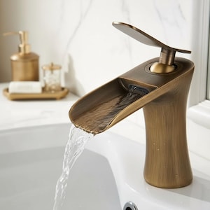 May include: A bronze-coloured bathroom tap with a waterfall spout. Water flows from the tap into a white sink. The tap has a decorative handle and is set against a white marble background. The tap is a stylish addition to any bathroom.