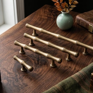 Może przedstawiać: Pięć mosiężnych uchwytów do szafek z bambusowym wzorem ułożonych na ciemnej drewnianej powierzchni. Uchwyty różnią się rozmiarem, największy ma około 25 cm długości. Obecny jest również mały wazon z suszonymi liśćmi i skórzana książka.