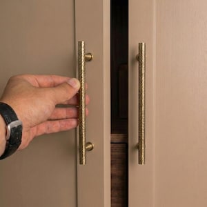 Puede incluir: Primer plano de una puerta de armario con dos tiradores verticales de latón texturizado. Los tiradores cilíndricos tienen un acabado martillado y están montados en un armario de tono neutro. La puerta del armario está parcialmente abierta.