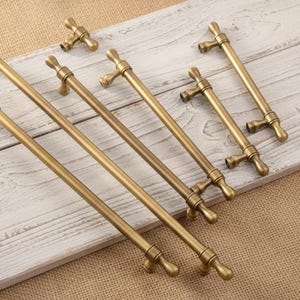 May include: A collection of antique brass cabinet pulls and knobs. The pulls are long and cylindrical with decorative ends, while the knobs are smaller with a similar design. They are arranged on a white-painted wooden surface.