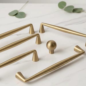 Peut inclure: Ensemble de quincaillerie de placard dorée, comprenant des poignées de barre et un bouton rond, disposé sur une surface en marbre blanc. La quincaillerie présente un design élégant et moderne, avec une finition mate. Les poignées varient en taille et en forme.