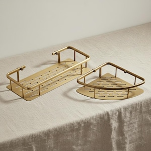May include: Two brass-coloured metal corner shelves. One is rectangular, the other is triangular. Both have a slotted base and a curved railing. The shelves are designed for bathroom or kitchen storage.