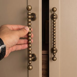 Peut inclure: Gros plan sur deux poignées de placard en laiton antique. Les poignées, avec un design perlé, sont montées sur une porte de placard marron clair. Une main ouvre une poignée.