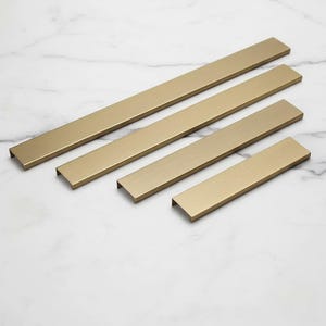 Pode incluir: Quatro puxadores dourados de diferentes comprimentos estão dispostos sobre uma superfície de mármore branco. Os puxadores têm um acabamento em metal escovado e um design simples e moderno. O puxador mais longo tem cerca de 30 cm.