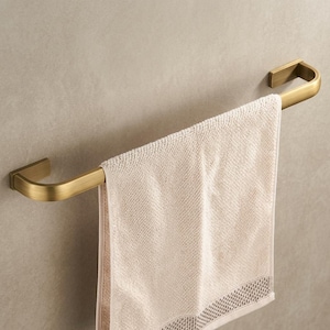 May include: A brass-colored towel bar with a curved design, mounted on a neutral-toned wall. A beige towel hangs from the bar, showcasing a textured weave and a decorative border.