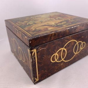 May include: A rectangular metal box with a brown faux-tortoiseshell finish and gold-coloured line accents. The lid features a printed landscape scene with figures, while the sides display a repeating pattern of interlocking gold-coloured circles.