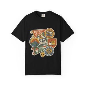 May include: Black t-shirt featuring a collection of colorful patches. Patches include a mountain scene, a camera, a cassette tape, a coffee cup, a mushroom, an alien, and a robot. Text on the patches reads "Adventure League", "Mixtape Memories", "Forage & Create", "Idea", and "Beep Boop Club".