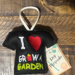 May include: Black felt ornament shaped like a t-shirt with the words "I ❤️ GROW A GARDEN" embroidered in white, red, and green. A tag reads "handmade by Amy Mercado" with yarn and crochet hook illustrations.