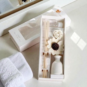 Könnte beinhalten: Ein weißes Aromatherapie-Diffusor-Set in einer Geschenkbox. Das Set enthält eine kleine weiße Keramikvase, eine klare Glasflasche, Holzstäbchen, eine dekorative weiße Blume und einen geflochtenen Ball. Die Box ist geöffnet und zeigt den Inhalt.