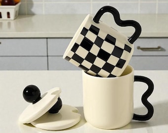 Puede incluir: Dos tazas de cerámica color crema, una con un patrón de cuadros blanco y negro, la otra lisa. Ambas tienen asas negras onduladas. Una tapa a juego con un pomo negro está al lado. Las tazas están apiladas.