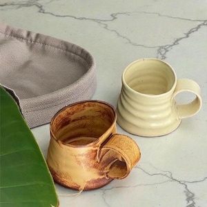 May include: Two handmade ceramic mugs. One is a light beige color with a ridged design, and the other is a brown and tan color with a unique handle. A gray fabric basket is in the background.