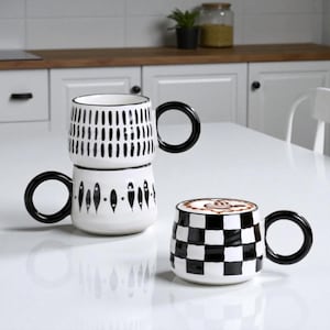 Può includere: Due tazze in ceramica bianca con manici neri. Una tazza ha un motivo a scacchi bianchi e neri, e l'altra ha un design impilato con motivi ovali e verticali neri. Le tazze sono su una superficie bianca.