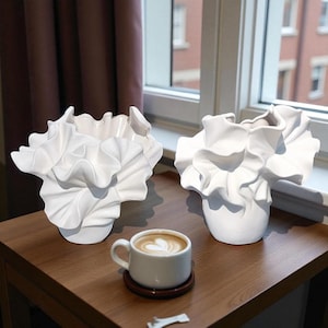 Könnte beinhalten: Zwei weiße Keramikvasen mit gerüschten, blütenblattartigen Designs stehen auf einem Holztisch. Eine kleine weiße Kaffeetasse mit herzförmigem Design im Schaum ruht auf einem Untersetzer. Die Vasen stehen in der Nähe eines Fensters.