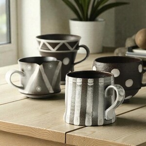 Puede incluir: Una colección de tazas de cerámica con diseños únicos. Las tazas presentan varios patrones, incluyendo rayas, lunares y formas geométricas. La paleta de colores incluye tonos marrones, blancos y grises. Las tazas están dispuestas sobre una superficie de madera.