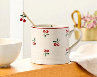 May include: A cream-colored ceramic mug with a red rim and handle, adorned with a repeating pattern of red cherries and green leaves. A small spoon with cherry details rests inside the mug. The mug is part of a kitchenware set.